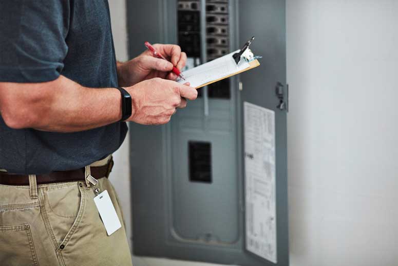 A-Phase Certified Electrician standing at electrical panel writing inspection report.