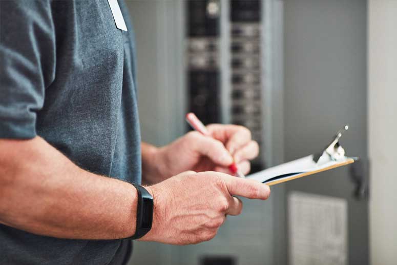 A-Phase Certified Electrician standing at electrical panel writing inspection report.